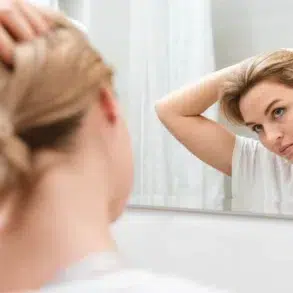 A woman in a white t-shirt uses a bathroom mirror to adjust her human hair topper for women.
