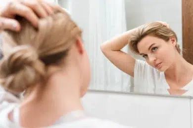 A woman in a white t-shirt uses a bathroom mirror to adjust her human hair topper for women.