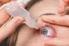 Close-up of a person applying eye drops to an irritated blue eye with visible redness. The hand gently holds the eyelid open, conveying caution and care.