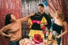 Three people holding costumes and props, standing in front of red and silver tinsel backdrops at a festive party or photo booth setup.