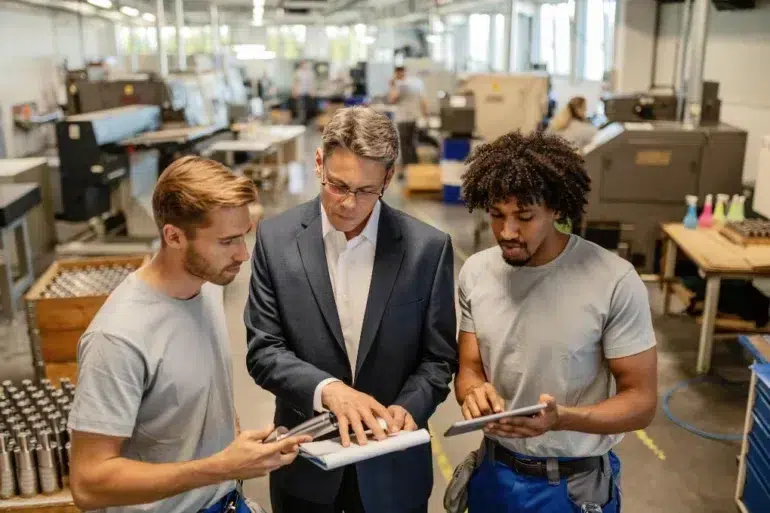 A manager in a suit discusses plans with two workers holding tablets in a modern factory. The environment is collaborative and focused.