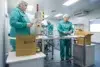 Workers in a clean room, wearing hairnets and green uniforms, perform industrial sorting and packaging at stainless steel tables.