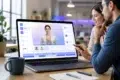 Two professionals review an AI-powered video editing interface on a laptop, showing a virtual presenter, timeline controls, and interactive training video elements in a modern office workspace.