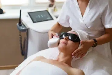A woman wearing protective goggles receives a laser facial treatment in a spa setting. The aesthetician, in white, focuses on her task in a calm atmosphere.