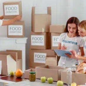Two women in volunteer shirts organize food donations, writing on a clipboard. A man stacks labeled boxes behind them. The mood is collaborative and positive.