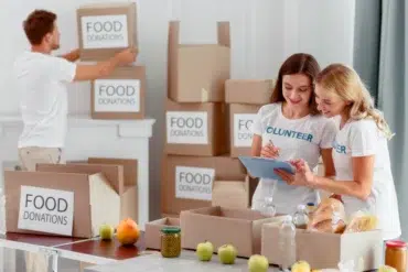Two women in volunteer shirts organize food donations, writing on a clipboard. A man stacks labeled boxes behind them. The mood is collaborative and positive.