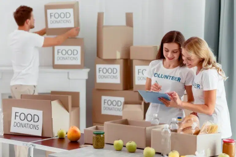 Two women in volunteer shirts organize food donations, writing on a clipboard. A man stacks labeled boxes behind them. The mood is collaborative and positive.