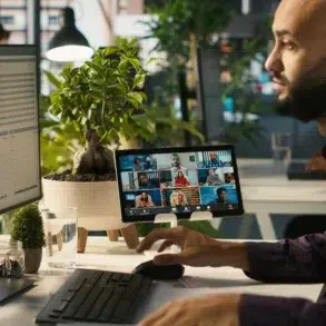 Man with a beard working at a desk with dual screens, participating in a video conference call in a modern office setting.