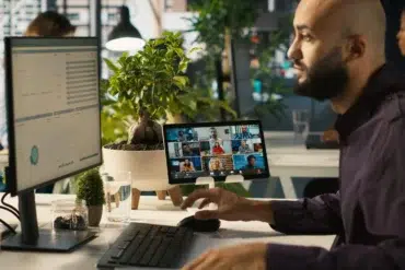 Man with a beard working at a desk with dual screens, participating in a video conference call in a modern office setting.