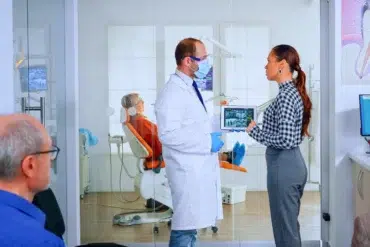 A dentist in a white coat discusses a dental X-ray on a tablet with a woman in a gingham shirt. An older person sits in a dental chair in the background.