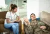 Healthcare professional listens to a man in military uniform lying on a couch during a therapy session at home.