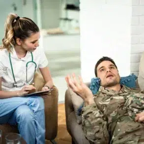 Healthcare professional listens to a man in military uniform lying on a couch during a therapy session at home.