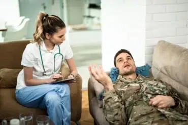 Healthcare professional listens to a man in military uniform lying on a couch during a therapy session at home.