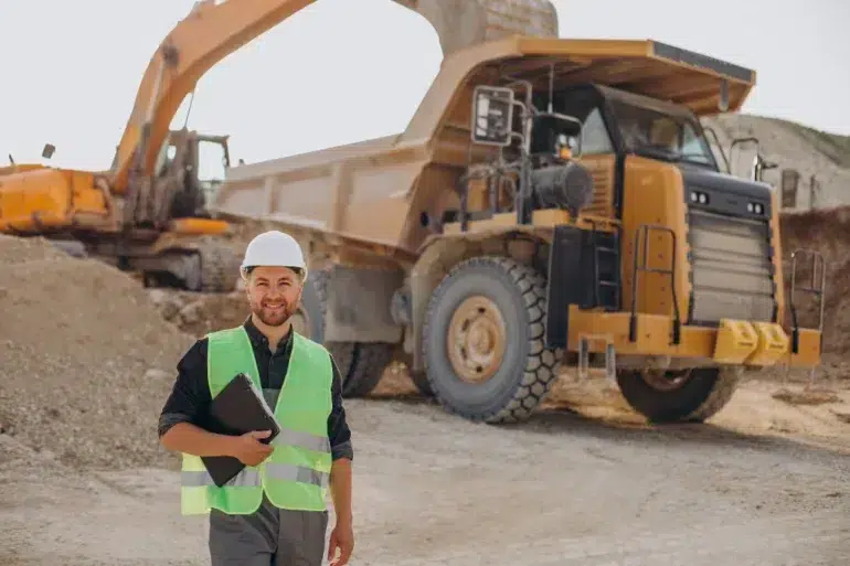 Man in a hard hat and reflective vest smiles while holding documents on a construction site. Large dump truck and excavator in the background.