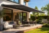 Modern patio with a retractable awning over a wooden deck, featuring a cozy sofa, dining table, and lush garden. The scene exudes relaxation.