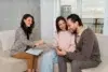 A couple sitting on a couch smiles as a woman with a clipboard discusses something with them. The atmosphere is friendly and supportive.