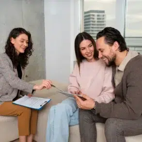 A couple sitting on a couch smiles as a woman with a clipboard discusses something with them. The atmosphere is friendly and supportive.