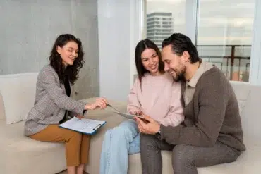 A couple sitting on a couch smiles as a woman with a clipboard discusses something with them. The atmosphere is friendly and supportive.