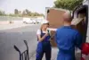 Two delivery workers in blue uniforms unload boxes from a van on a street. The woman holds a clipboard and talks to a male colleague, conveying teamwork.