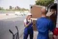 Two delivery workers in blue uniforms unload boxes from a van on a street. The woman holds a clipboard and talks to a male colleague, conveying teamwork.