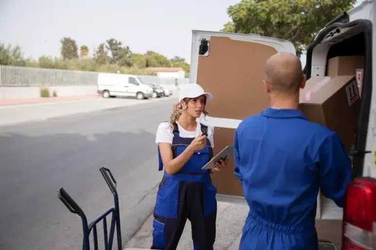 Two delivery workers in blue uniforms unload boxes from a van on a street. The woman holds a clipboard and talks to a male colleague, conveying teamwork.