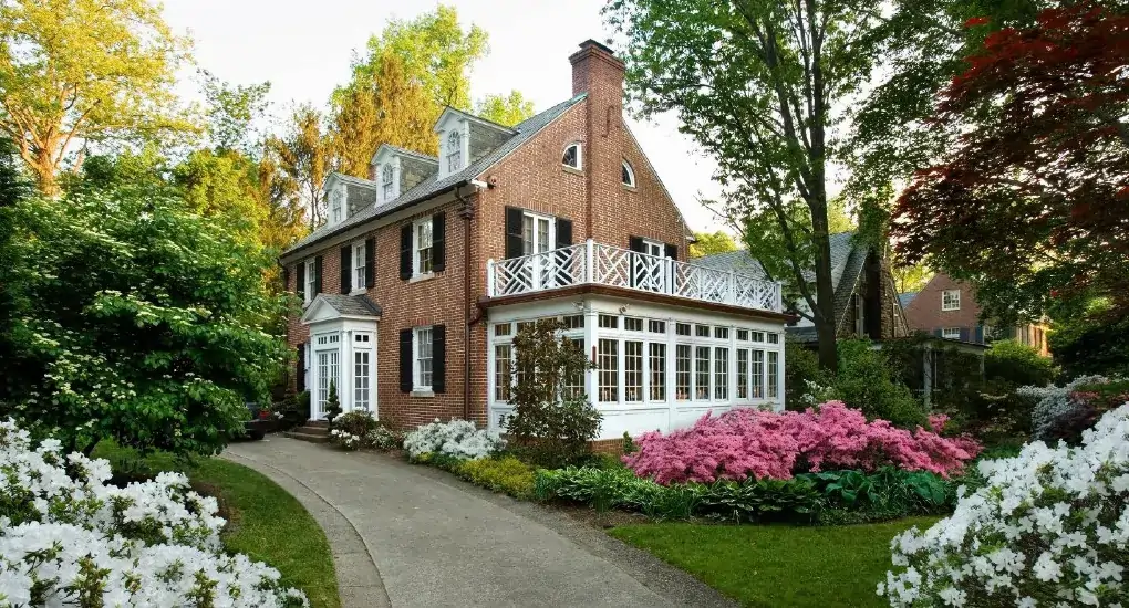 Brick colonial-style house with white-trimmed windows, a curved driveway, and colorful flowering bushes in a lush garden.