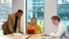 Three colleagues in a modern office discuss work at a table. A woman in a yellow blazer holds a red folder, while a man in a suit leans over, and another types on a laptop. The mood is collaborative.
