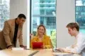 Three colleagues in a modern office discuss work at a table. A woman in a yellow blazer holds a red folder, while a man in a suit leans over, and another types on a laptop. The mood is collaborative.