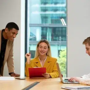 Three colleagues in a modern office discuss work at a table. A woman in a yellow blazer holds a red folder, while a man in a suit leans over, and another types on a laptop. The mood is collaborative.