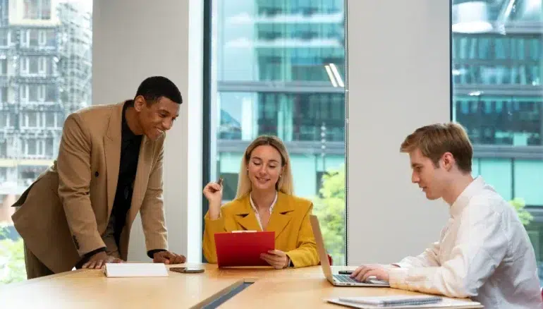 Three colleagues in a modern office discuss work at a table. A woman in a yellow blazer holds a red folder, while a man in a suit leans over, and another types on a laptop. The mood is collaborative.