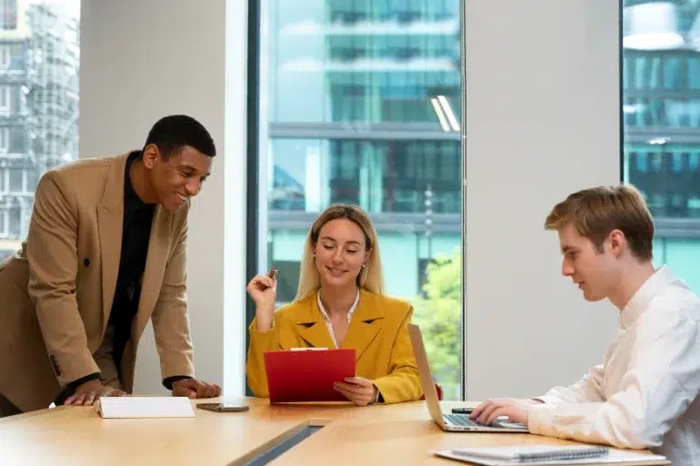 Three colleagues in a modern office discuss work at a table. A woman in a yellow blazer holds a red folder, while a man in a suit leans over, and another types on a laptop. The mood is collaborative.