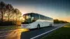 A white coach bus travels on a highway at sunrise, with sunlight reflecting off its windshield. Trees line the road, and the sky glows orange, creating a serene scene.