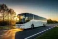 A white coach bus travels on a highway at sunrise, with sunlight reflecting off its windshield. Trees line the road, and the sky glows orange, creating a serene scene.