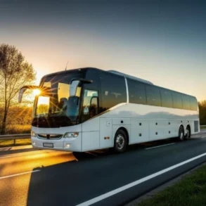 A white coach bus travels on a highway at sunrise, with sunlight reflecting off its windshield. Trees line the road, and the sky glows orange, creating a serene scene.