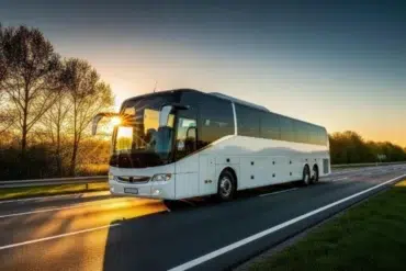 A white coach bus travels on a highway at sunrise, with sunlight reflecting off its windshield. Trees line the road, and the sky glows orange, creating a serene scene.