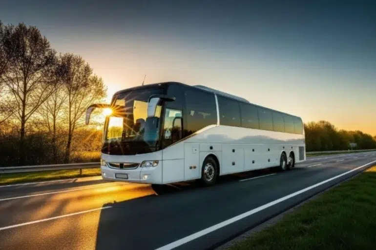 A white coach bus travels on a highway at sunrise, with sunlight reflecting off its windshield. Trees line the road, and the sky glows orange, creating a serene scene.