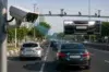 A high-tech surveillance camera mounted on a pole captures traffic on a multi-lane road, demonstrating a Number Plate Recognition System that identifies and highlights license plates on moving vehicles.