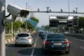 A high-tech surveillance camera mounted on a pole captures traffic on a multi-lane road, demonstrating a Number Plate Recognition System that identifies and highlights license plates on moving vehicles.