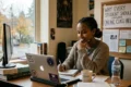 A woman sits at a desk smiling while looking at her laptop. Books, a mug, and a water bottle are nearby. A sign reads "Why Every Student Should Consider Online Class Help."