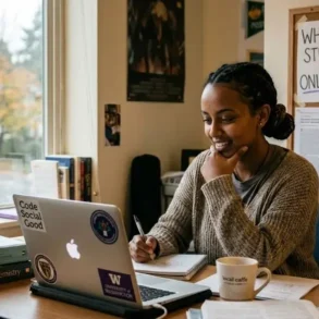 A woman sits at a desk smiling while looking at her laptop. Books, a mug, and a water bottle are nearby. A sign reads "Why Every Student Should Consider Online Class Help."
