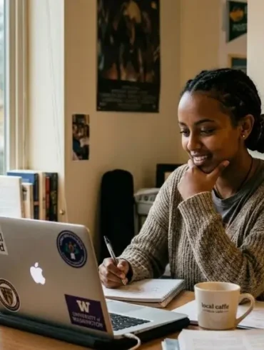 A woman sits at a desk smiling while looking at her laptop. Books, a mug, and a water bottle are nearby. A sign reads "Why Every Student Should Consider Online Class Help."