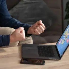 A person in a blue shirt clenches their fists in excitement while sitting in front of a laptop displaying a colorful bingo game screen.