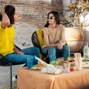 A man and woman sit outdoors, chatting beside a table with food and drinks. They appear relaxed and are surrounded by a rustic brick wall.
