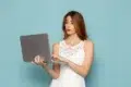Woman in a white lace dress holds and types on a laptop against a light blue background, appearing focused and engaged.