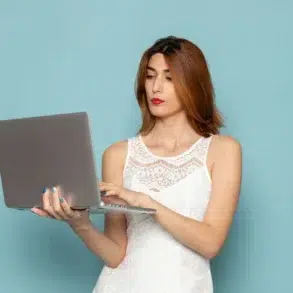 Woman in a white lace dress holds and types on a laptop against a light blue background, appearing focused and engaged.
