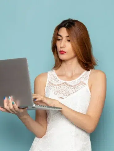 Woman in a white lace dress holds and types on a laptop against a light blue background, appearing focused and engaged.