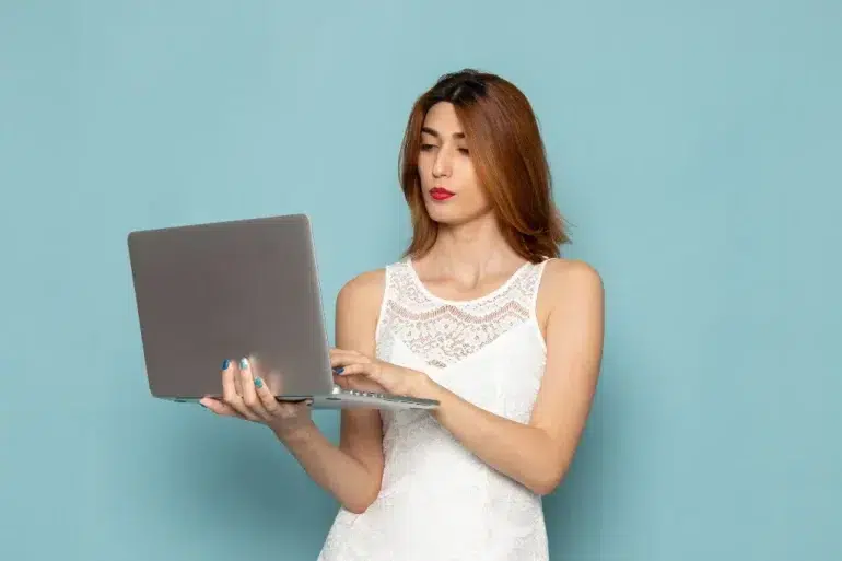 Woman in a white lace dress holds and types on a laptop against a light blue background, appearing focused and engaged.