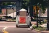 White utility vehicle with a sign "Litter Crew Ahead, This Vehicle Makes Frequent Stops" parked on a street under a bridge near trees and road signs.