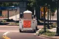 White utility vehicle with a sign "Litter Crew Ahead, This Vehicle Makes Frequent Stops" parked on a street under a bridge near trees and road signs.