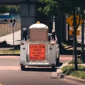 White utility vehicle with a sign "Litter Crew Ahead, This Vehicle Makes Frequent Stops" parked on a street under a bridge near trees and road signs.
